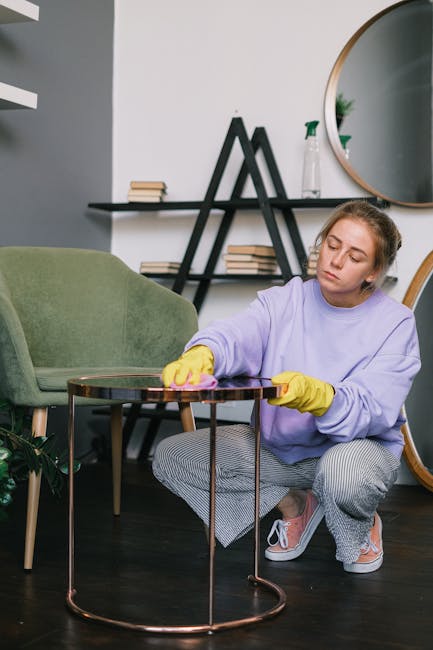 A person wearing a black leather jacket is vacuuming a patterned yellow and white cushion placed on a dark grey fabric sofa. The vacuum cleaner hose is black and the person’s hand is gripping the ha