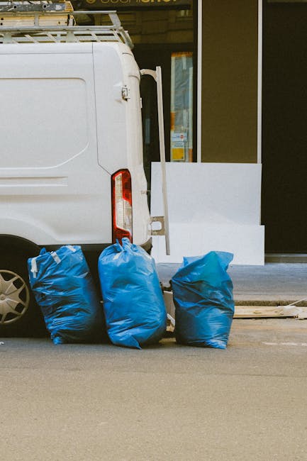 A black commercial cleaning van parked in an outdoor parking area beneath a concrete structure, with its rear door open revealing shelving units filled with cleaning tools and supplies, including spra