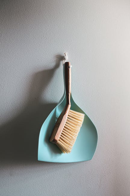 A light blue dustpan hanging on a white wall with a wooden-handled broom resting inside it. The broom has beige bristles and is positioned vertically within the dustpan. The scene is well-lit, showcas