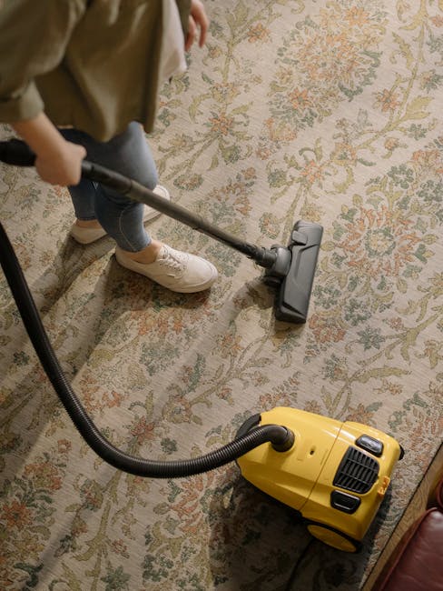 Close-up of a person vacuuming a patterned area rug with a black and gray vacuum cleaner in a well-lit room. The floor beneath the rug is wooden, and the person is wearing white sneakers and dark pant